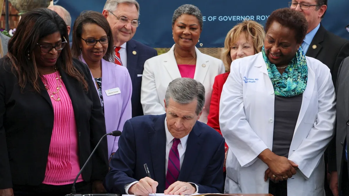 North Carolina governor sit at a table and signs a Medicaid expansion bill. There are numerous people standing to both sides of him.