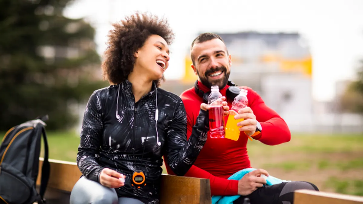 Two young athletes in sports clothing putting their energy drink bottles together while sitting on a bench in a public park
