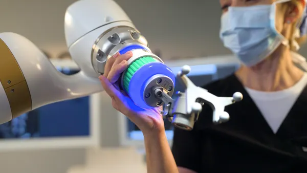 A medical professional in a face mask is standing next to the arm of a surgical robot.