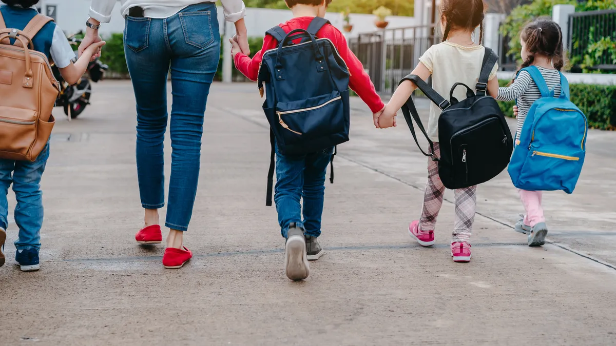 An adult is holding hands with children carrying backpacks. Their backs are toward to the camera