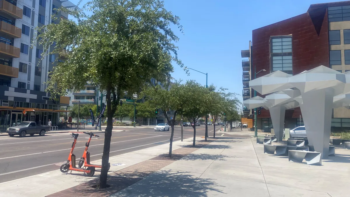 A sidewalk along a street is lined with several trees. Deeper into the sidewalk are benches and large built structures casting shadows.