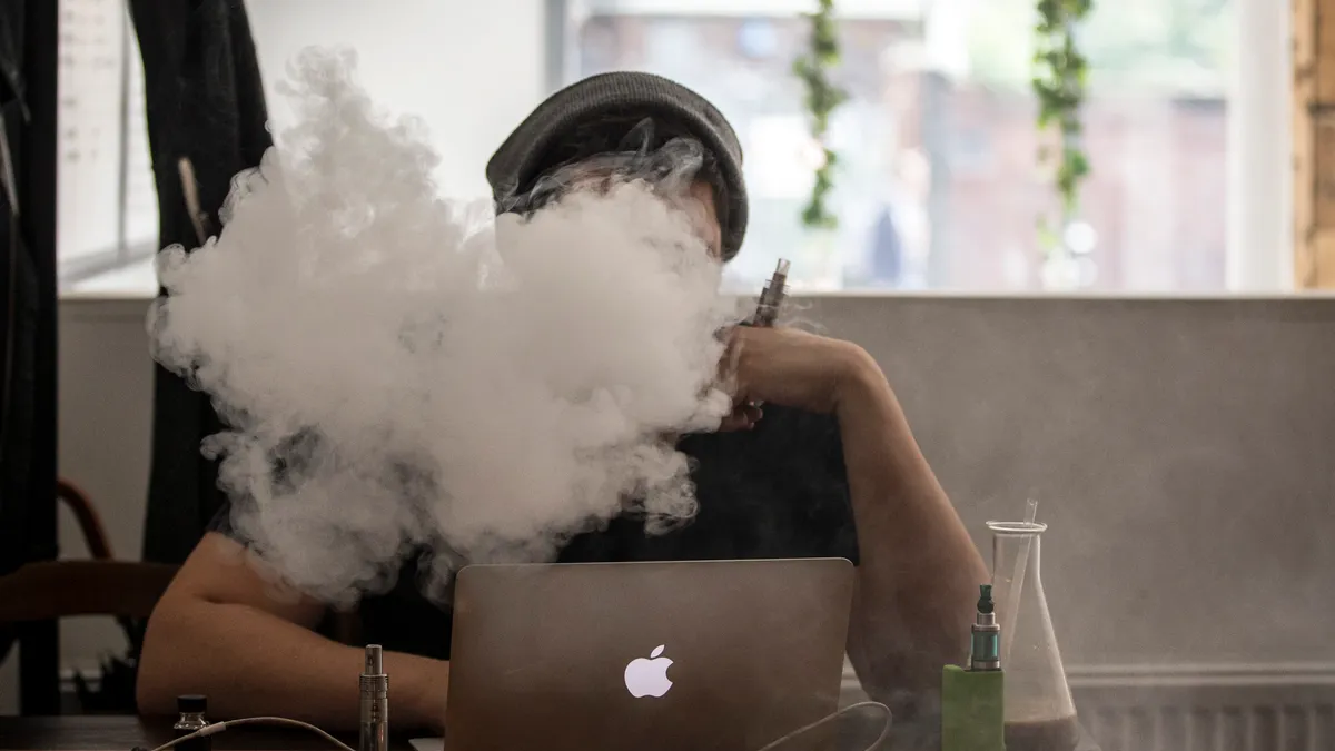 A person sits at a desk with an open laptop in front of them. Their head is hidden by smoke and the hand is holding a vaping device.