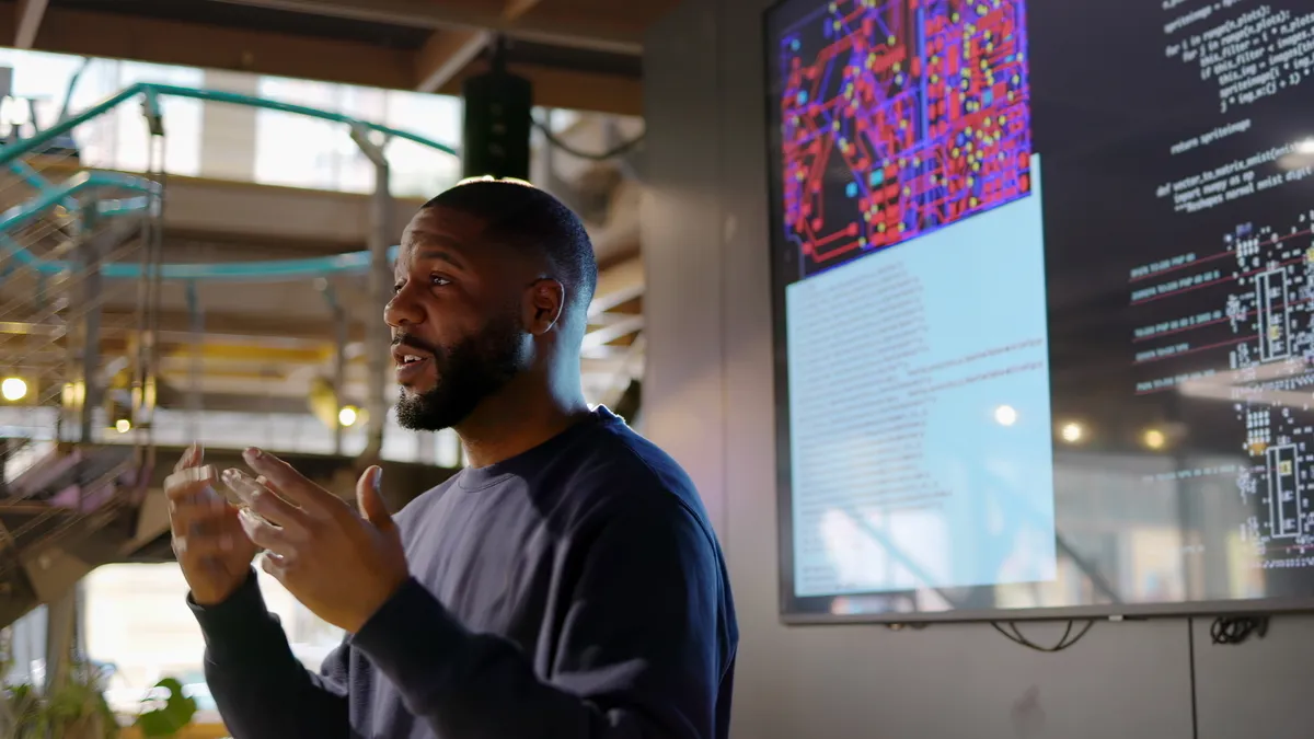 A Black man stands at a screen, teaching a seminar on Python coding.