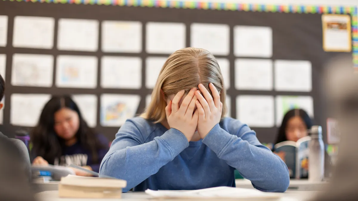 A student sitting at a desk in a classroom as their hands over their face.