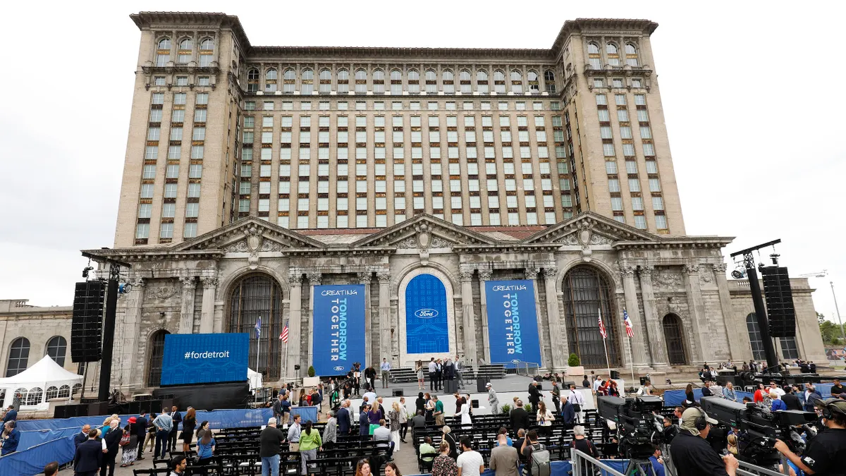 A crowd of people gather on the front steps of an older multistory building decorated with blue banners.