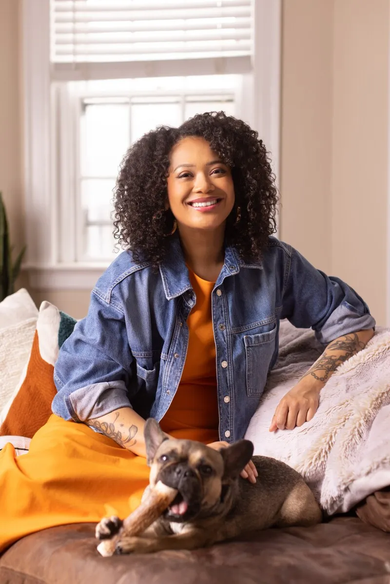 A woman in a jean jacket relaxes on a couch beside a French bulldog chewing on a bone.