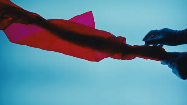 Against a blue background, a hand in a rubber glove holds onto the edge of a red transparent sheet of Lunaform material as it floats away.