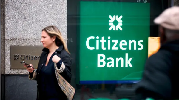 A Citizens Bank illuminated signage is framed between two walking pedestrians on the street. 