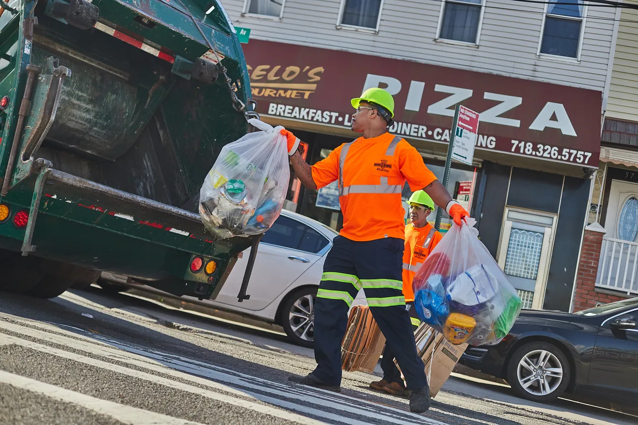 Two workers in orange shirts and yellow hard hats collect recyclables to throw into a truck, in front of a pizza restaurant in New York City