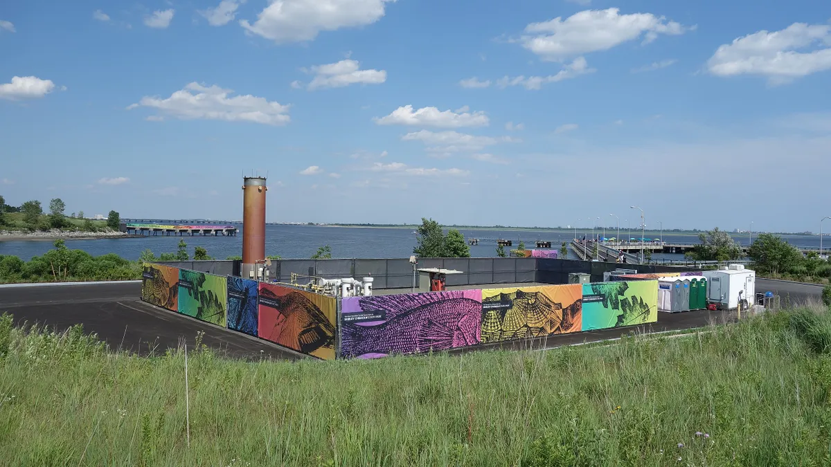 A brown cylindrical stack rises behind some decorated fencing on a grassy hill. A body of water is visible in the background.