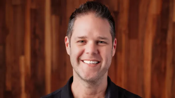 An image of a person wearing a black polo shirt smiling in front of a wood paneled background.