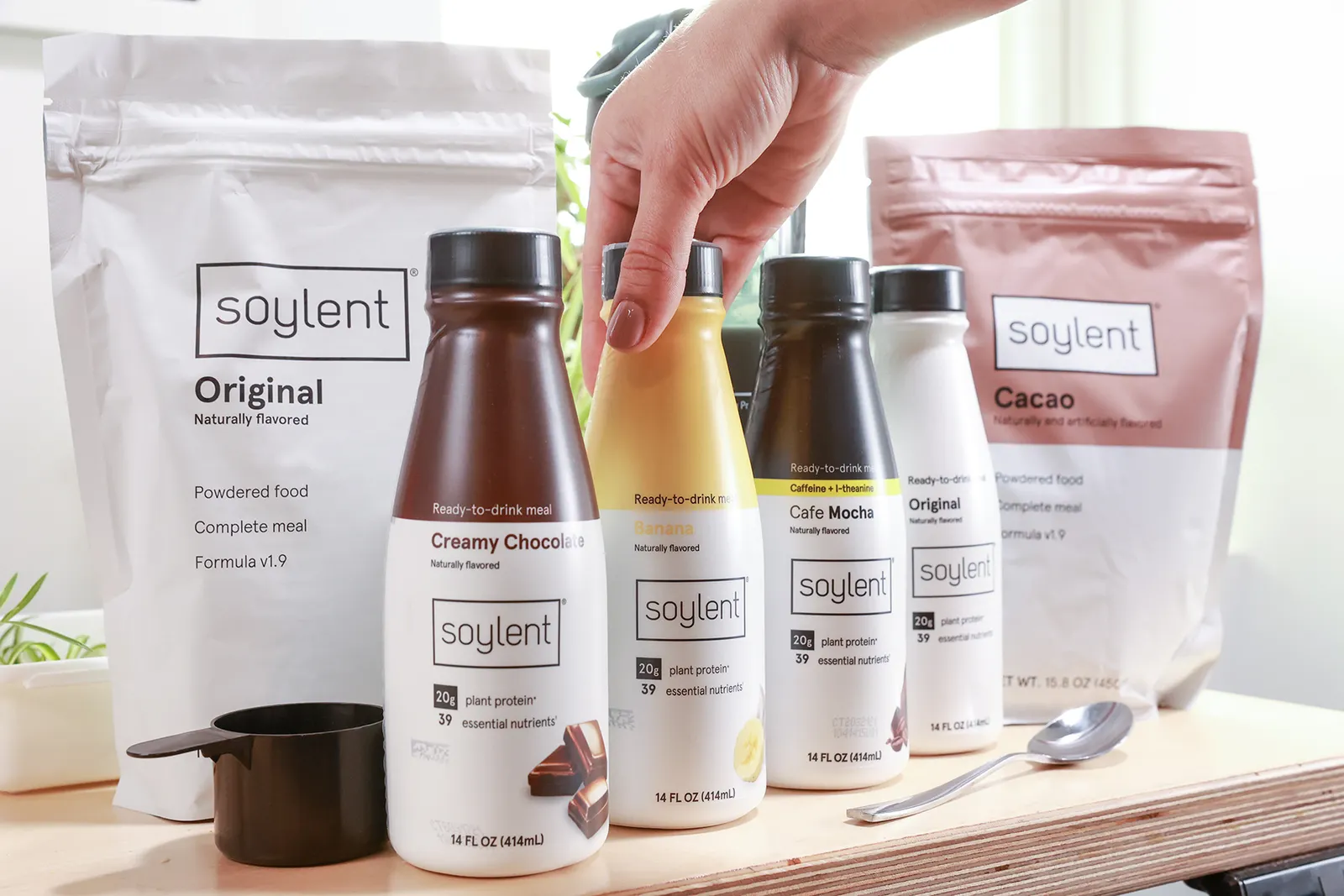 Two pouches of Soylent protein powder and three four different bottles of Soylent beverages sit on a table with a measuring cup and a spoon. A hand touches one of the bottles.