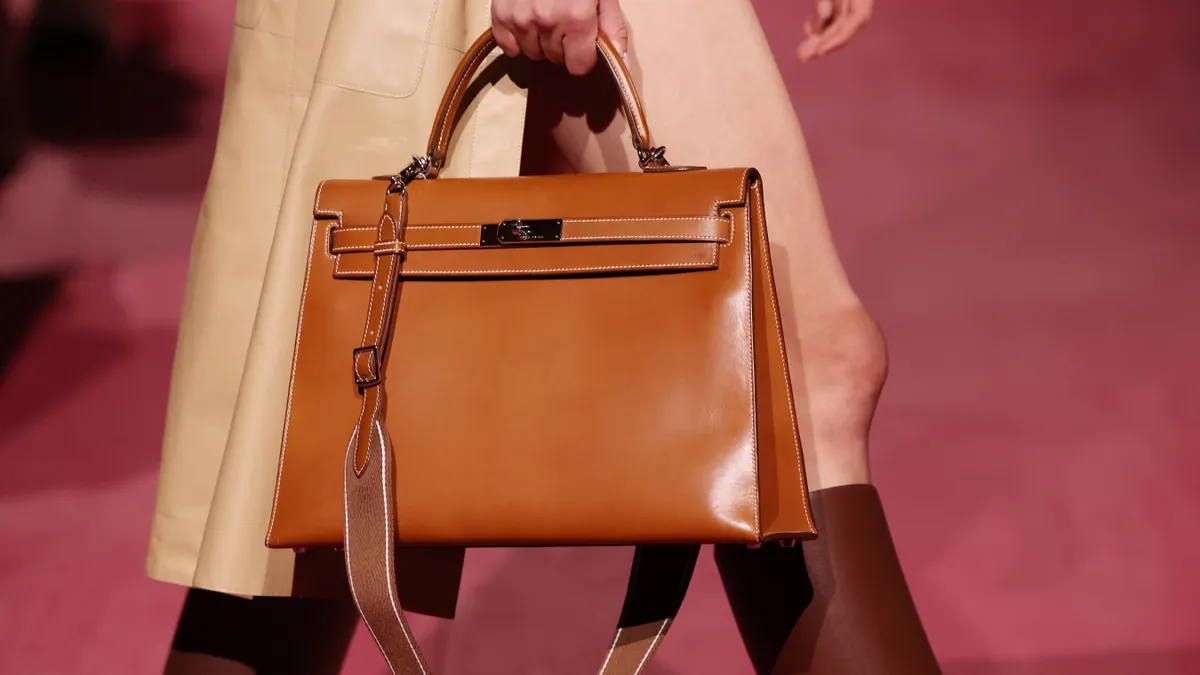 A model carries a brown leather Hermès Kelly bag while walking down a runway.