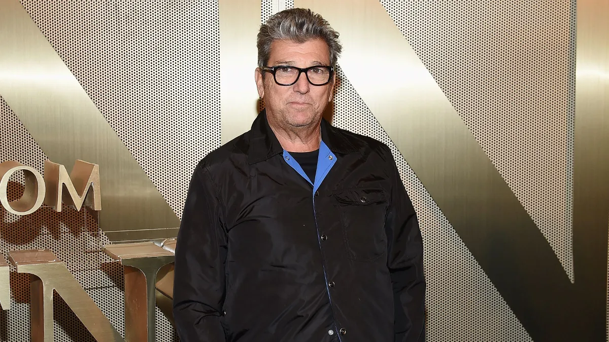 A person with gray hair and glasses wears a black jacket and stands in front of a gold wall looking at the camera.