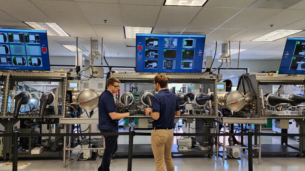 Two people in blue polo shirts and protective goggles in front of manufacturing machinery.