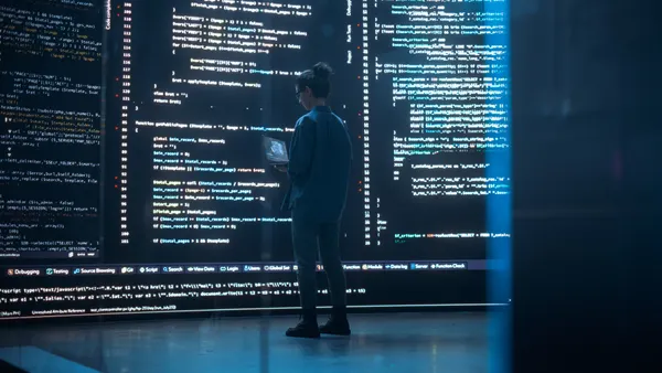a technologist holds a laptop, facing a wall of code projected on a wall
