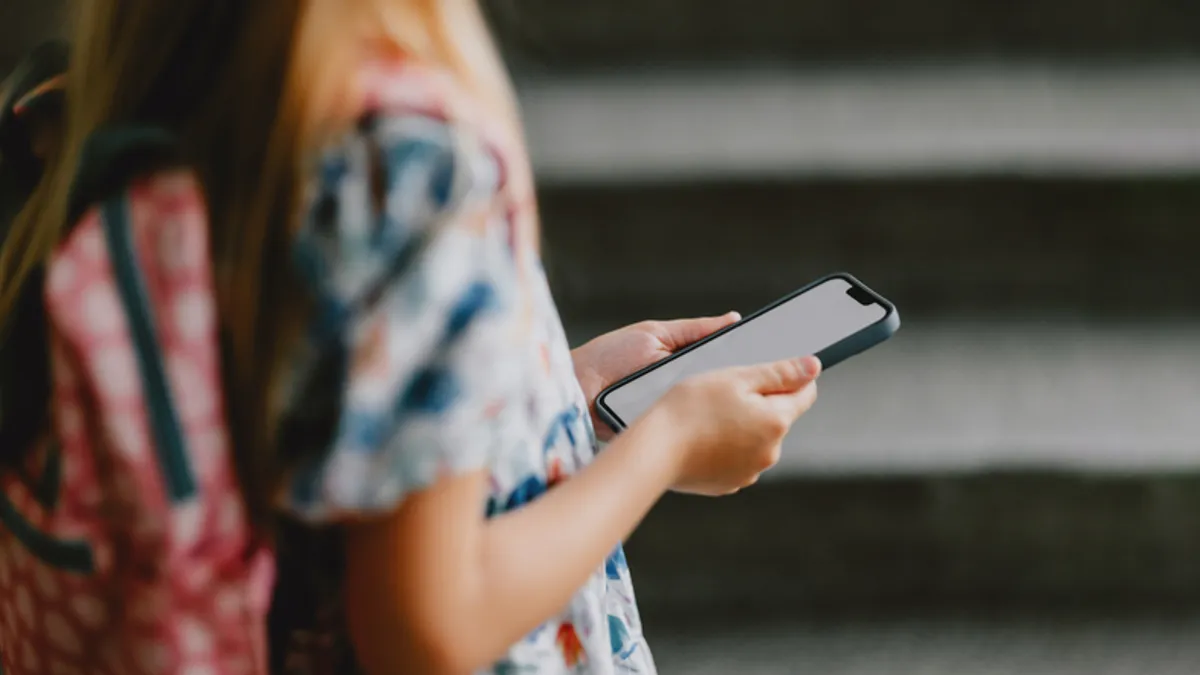 A student with a backpack is shown from the side from the shoulders down, In their hands is a cellphone