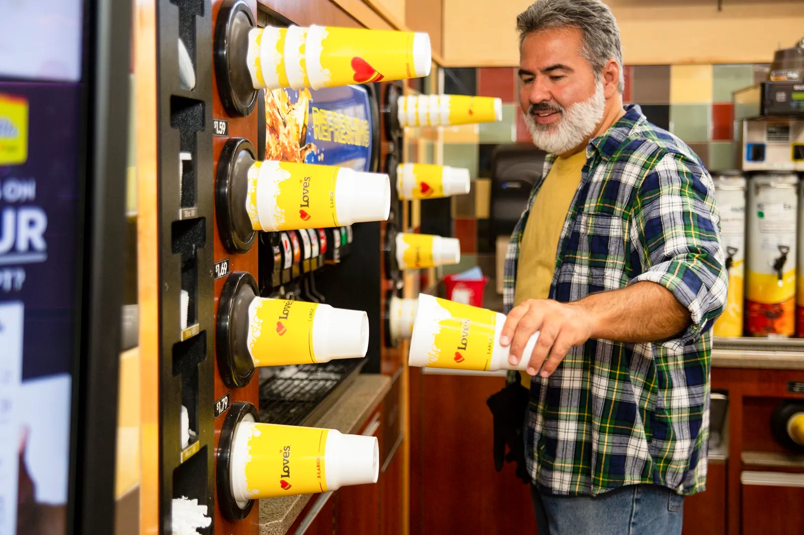 A photo of a person taking a cup from a stack of plastic cups on the wall. the cups are yellow and say &quot;Love&#x27;s.&quot; There is a soda dispenser with multiple flavors in the wall next to the cups.