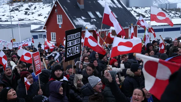 People wave the Greenland flag and hold signs during a protest. The signs read "Greenland is not for sale!"
