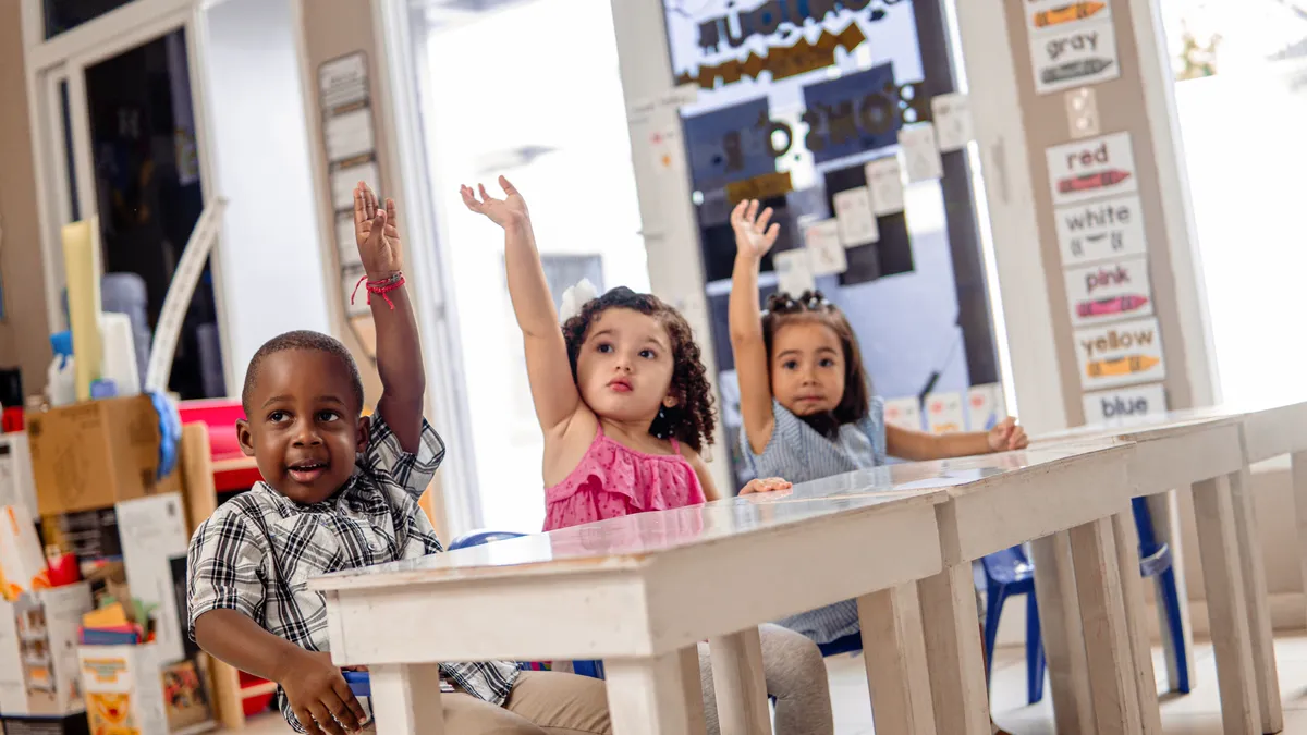 Three young students sit at a desk and have their hands raised