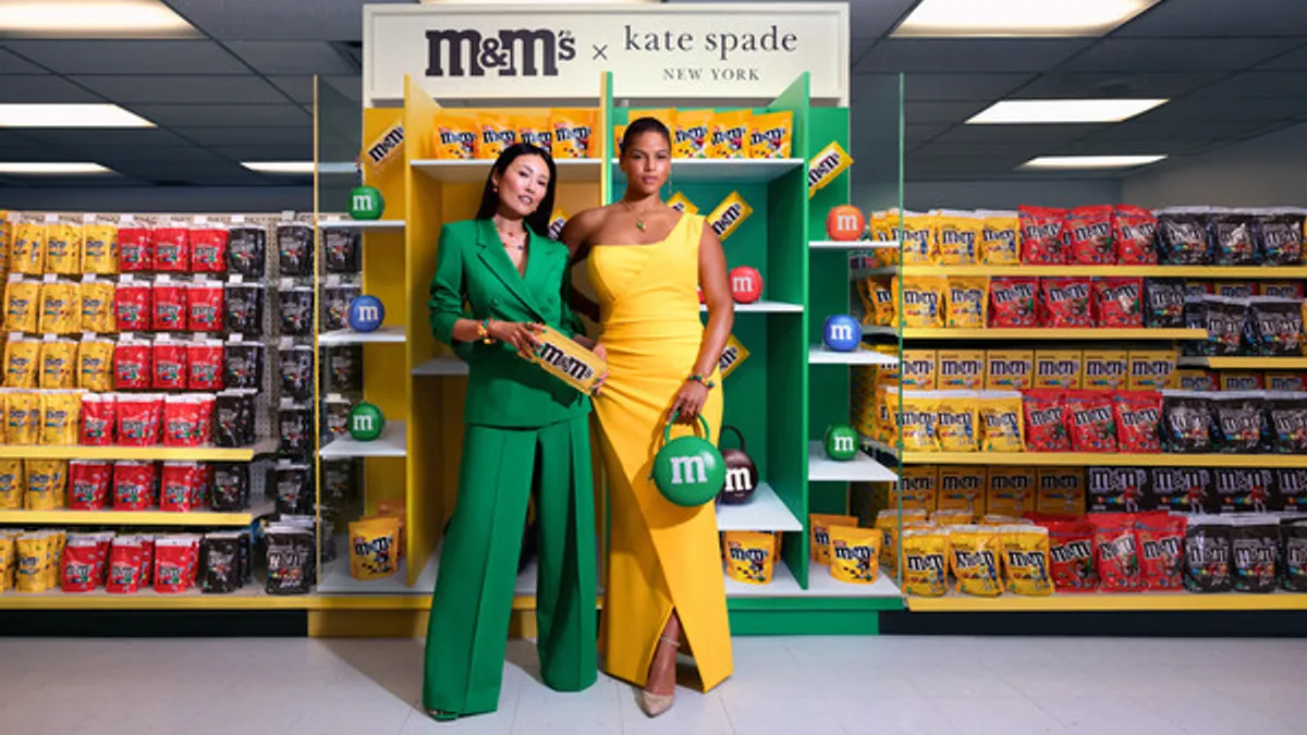 Two models holding bags from the collaboration collection in an M&M candy aisle of a store.