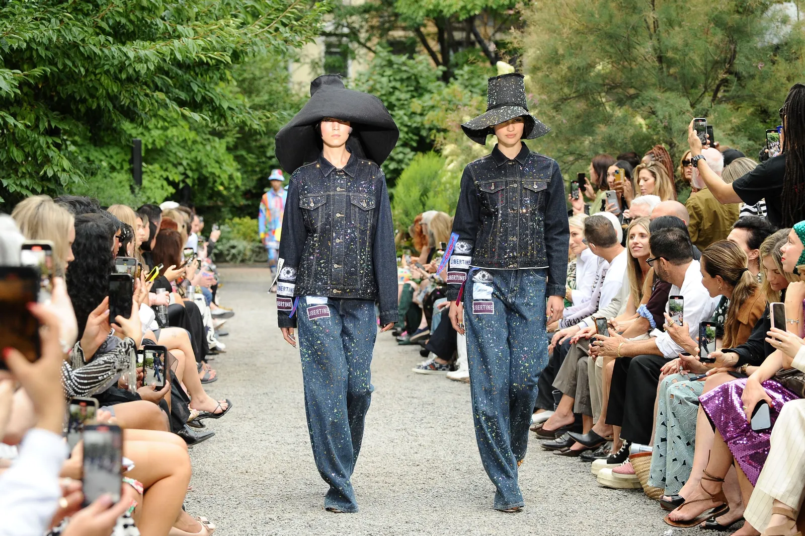 Two models in denim outfits walk side by side down a grassy runway.