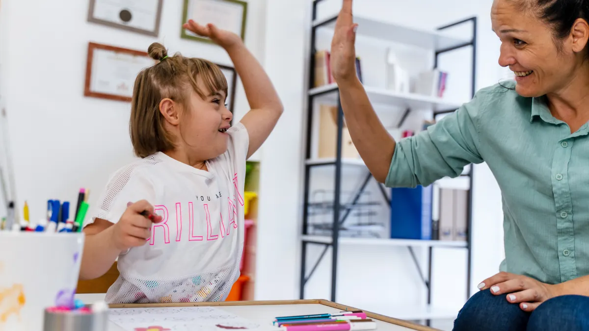A student and teacher are sitting at a table in a classroom. The student is holding a Crayon and is high fiving the teacher.
