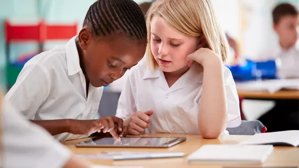 Two students are sitting next to each other at a desk in a classroom and are looking at a tablet