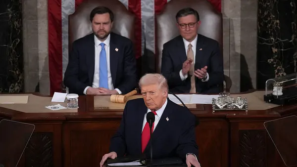 President Trump, wearing a red tie and a glower, is flanked by VP JD Vance and House Speaker Mike Johnson behind a podium.