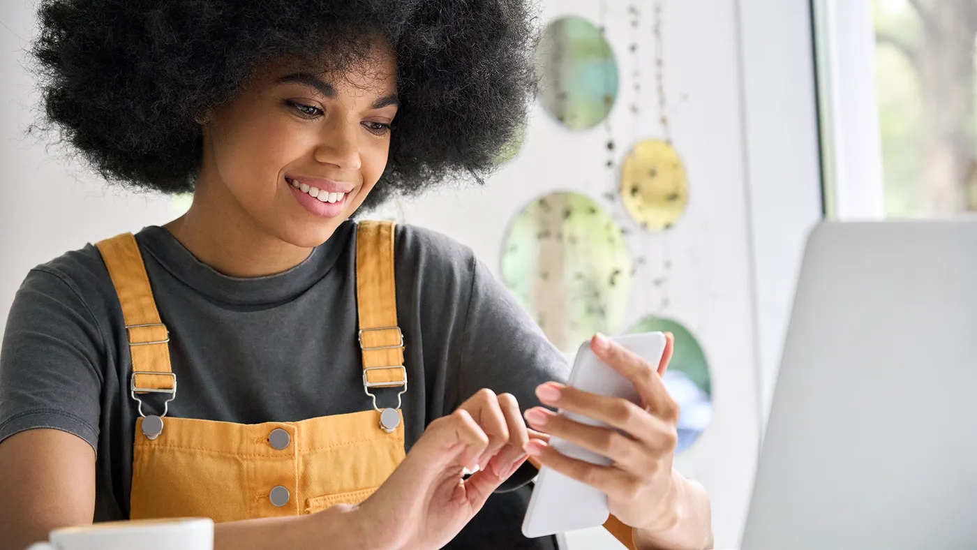 Woman with curly hair wearing a gray shirt and yellow overalls, sitting at a desk with a laptop and coffee cup.