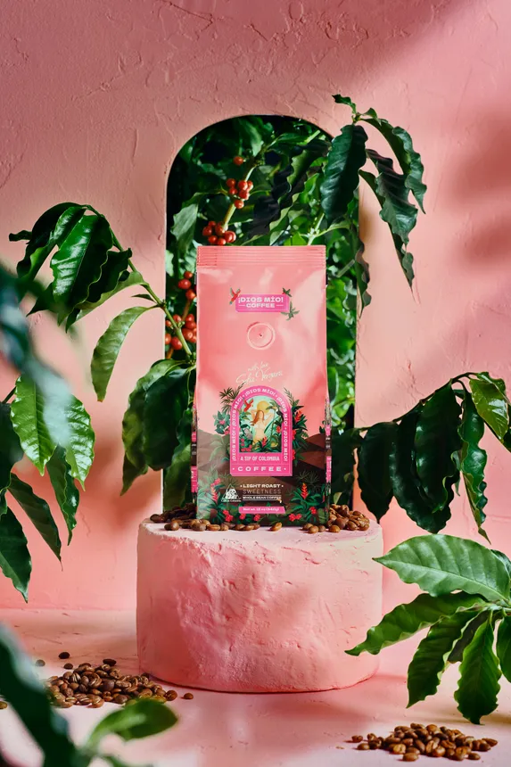 A bag of light roast beans from the ¡DÍOS MIO! COFFEE brand, surrounded by green plants with a pink background.