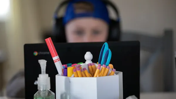 A young student wearing headphones works on a laptop behind a focused image of school supplies and a bottle of hand sanitizer.