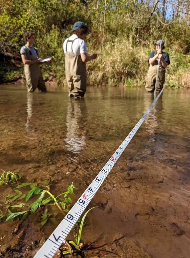 Students in coveralls standing in a stream with tape measure in foreground.