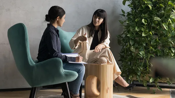 Two employees sitting down and talking next to a green plant.