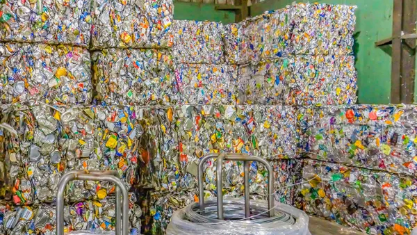 Stacks of colorful crushed can bales sit in a warehouse. There are two metal wire spools in front of them. 