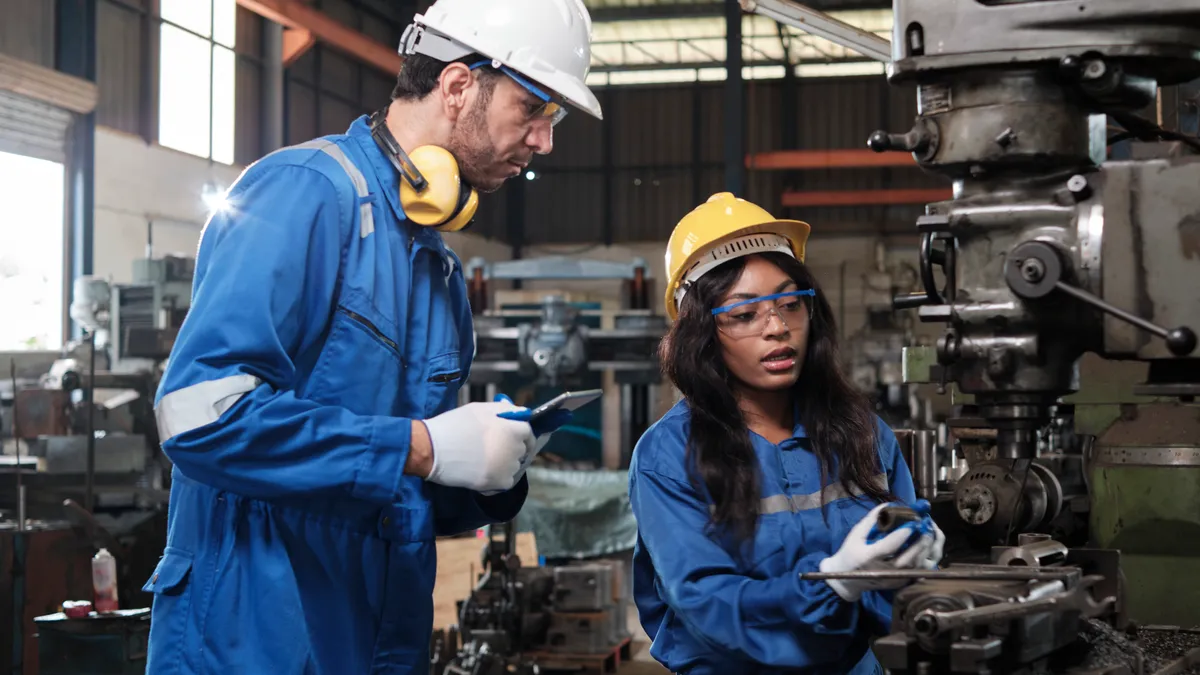 Two industrial workers in protective and safety uniforms and hardhats working with metalwork machines in a manufacturing factory.