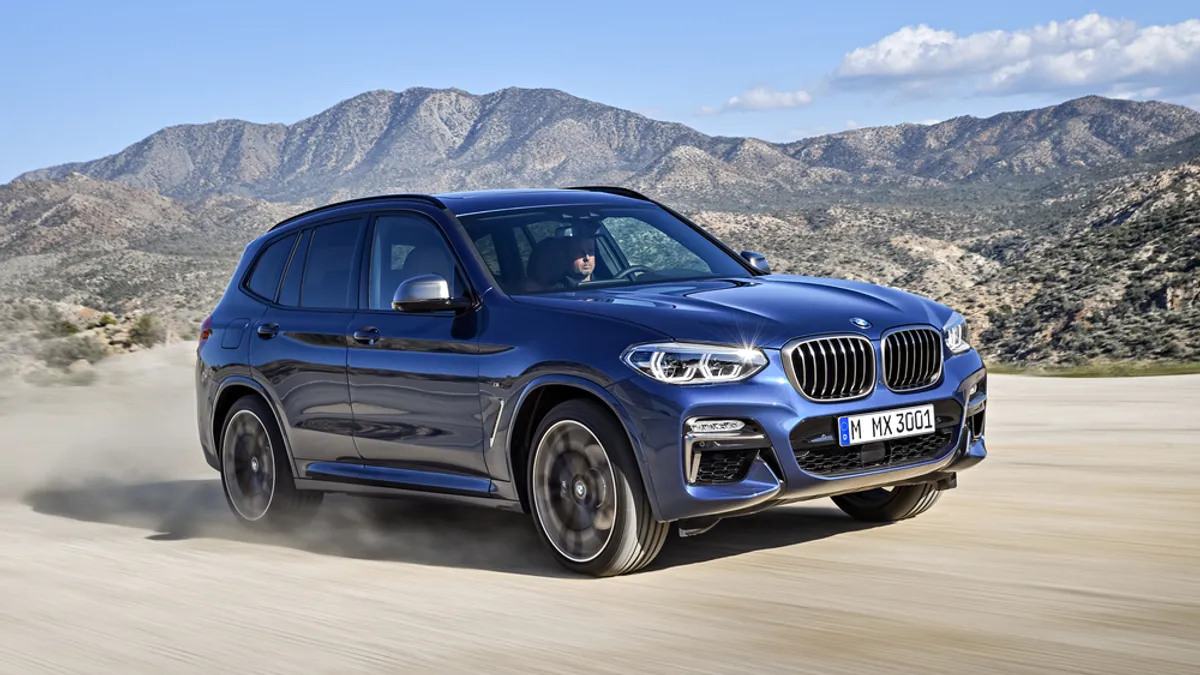 A Blue 2018 BMW X3 SUV pictured driving on an unpaved road with mountains in the background.