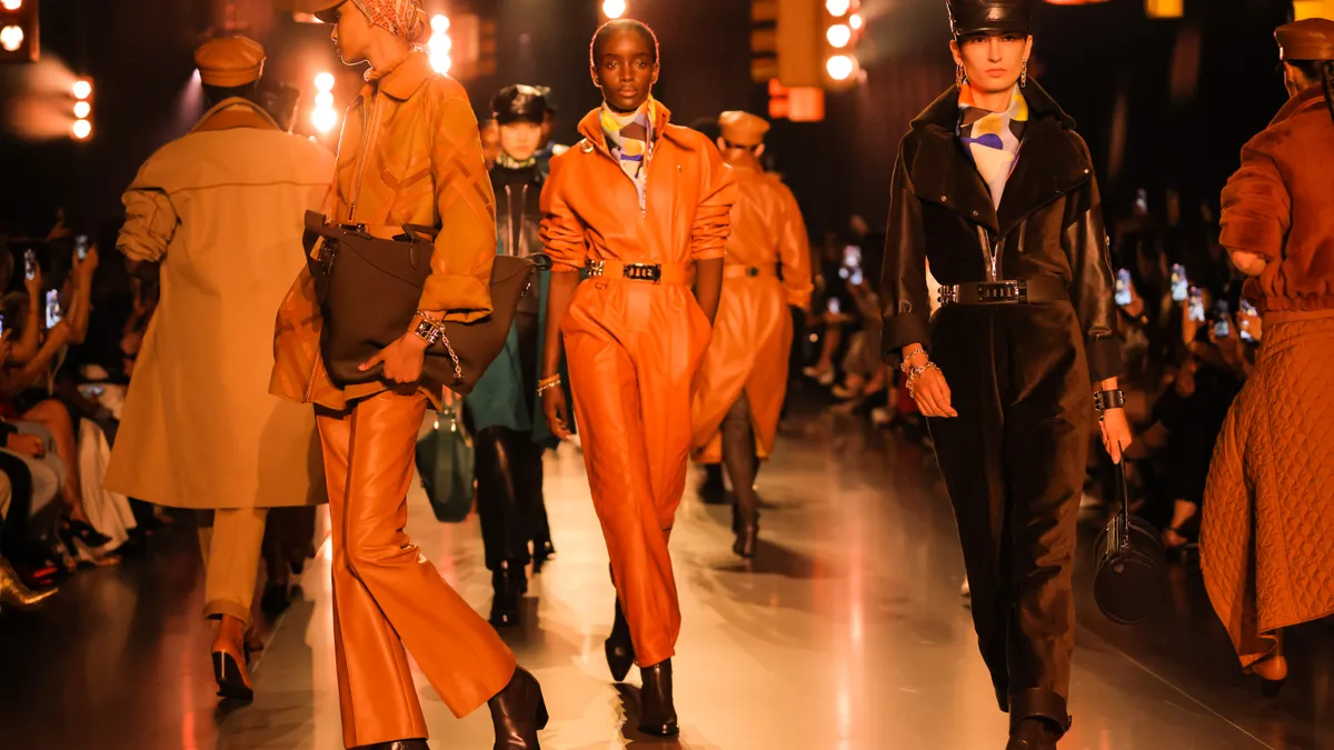 Models in orange outfits walk on a runway.