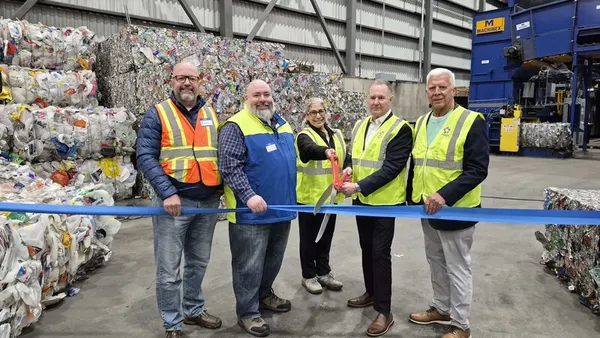 Workers hold large scissors to cut a ribbon inside a recycling facility. Bales of recycled plastics can be seen in the background.