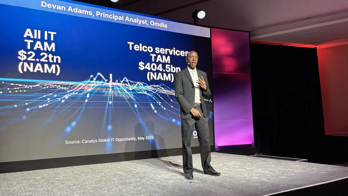 A man in a suit stands on a stage in front of a blue presentation screen. The screen shows his name, "Devan Adams, Principal Analyst, Omdia" and statistics about IT spending.