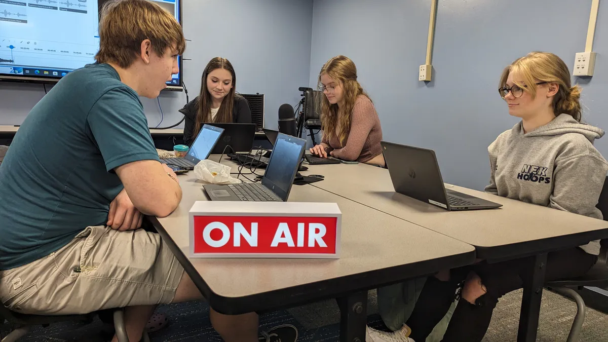 Four high school students sit around a desk with open laptops in a classroom. A sign on the desk reads "On Air."