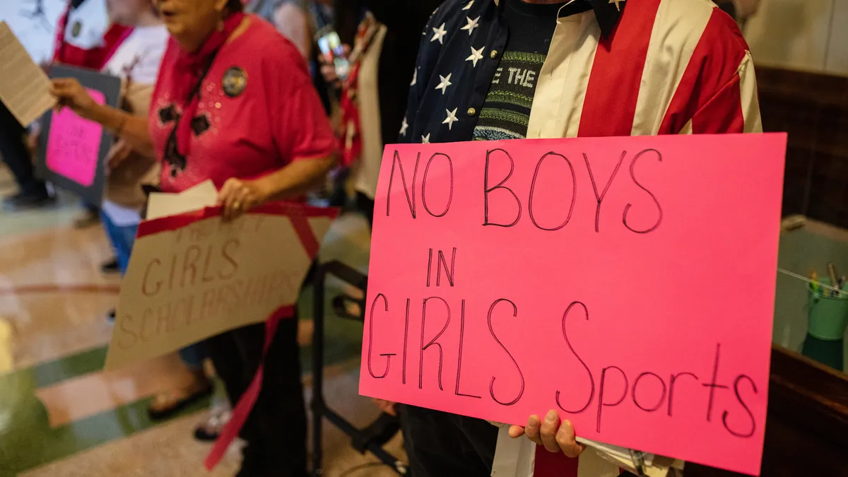 A demonstrator wearing an American flag shirt holds a protest sign saying "No Boys In Girls Sports"