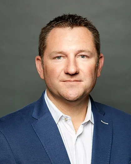 A white man wearing a blue suit jacket and a white casual shirt looks into the camera. When will executives start wearing neckties again?