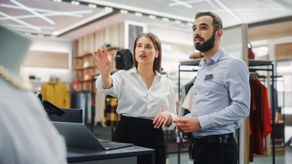 Clothing Store: Businesswoman Uses Laptop Computer, Talks to Visual Merchandising Specialist, Collaborate To Create Stylish Collection. Business Owner's Fashion Shop: Sales Manager Talks to Designer