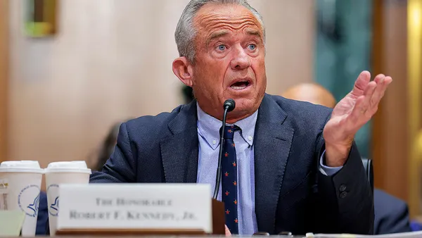 Person wearing a suit, collared shirt and tie speaking into a microphone behind a placcard