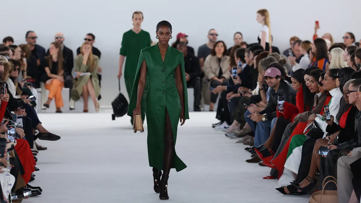 A model in a green outfit walks down a white runway as spectators sit watching from the side.