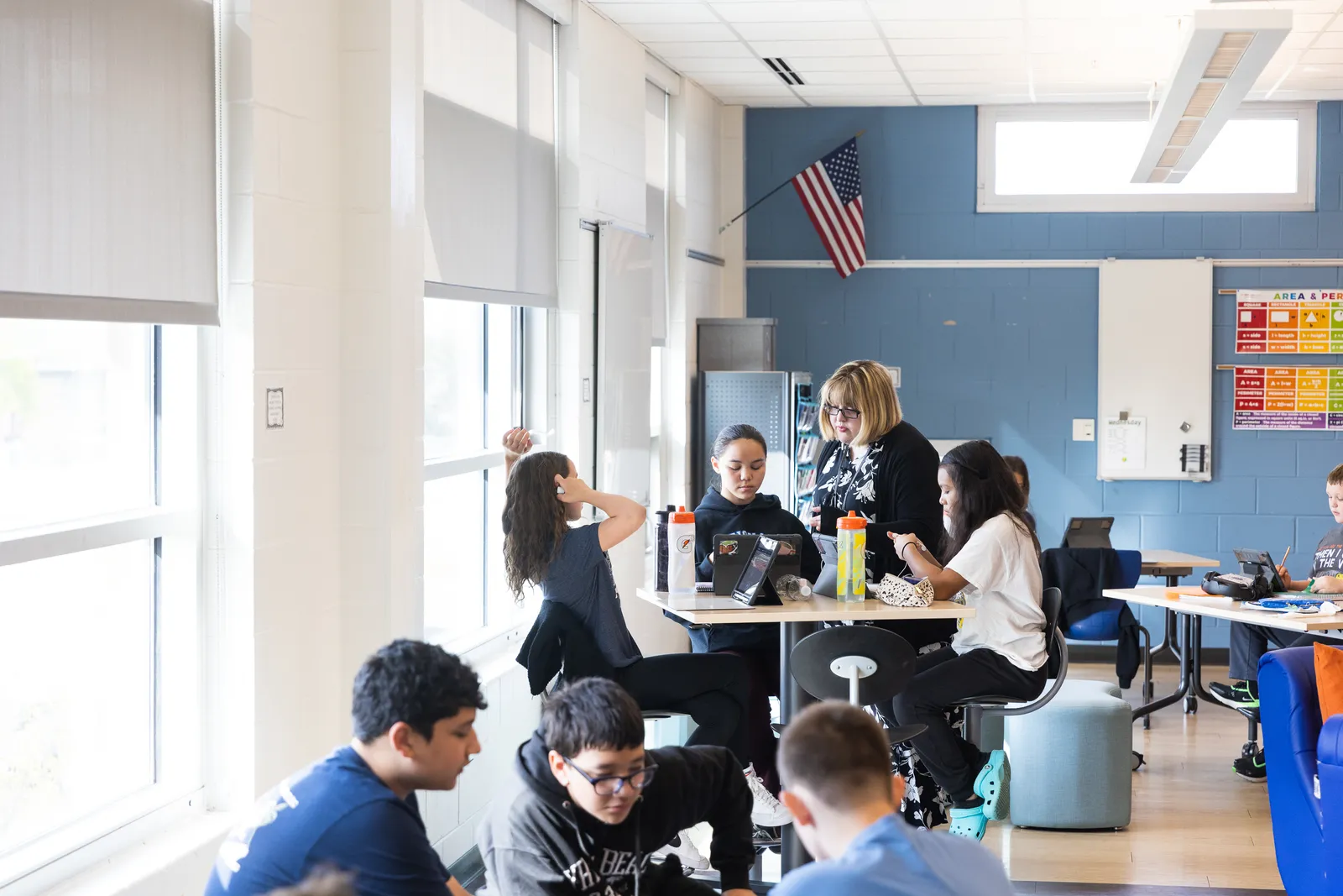 Students in a classroom are working in small groups at tables. An adult is working with one group of  three students.