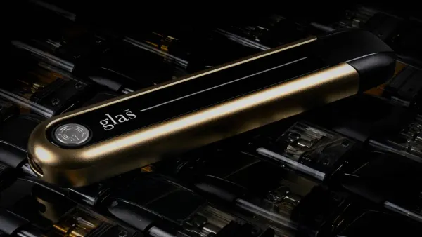 A vaping device lies on a bed of vaping liquid pods. The device is gold and black with a small silver seal and says "Glas" on one end.