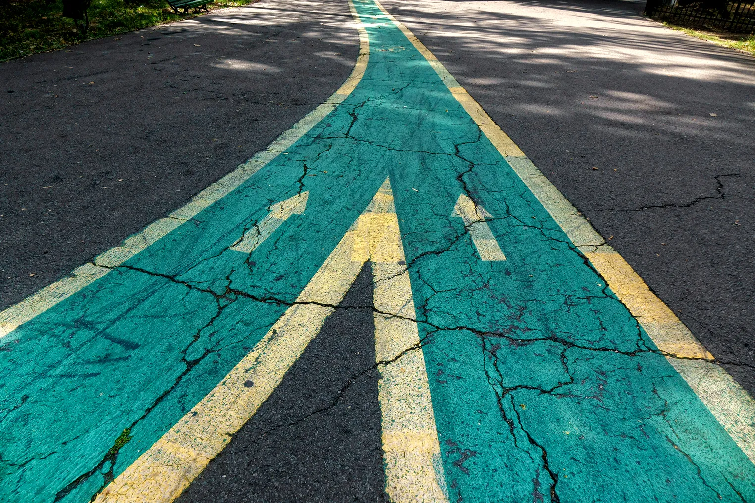 Two green roads, bordered by yellow paint, converge on an asphalt road with arrows pointing forward away from the frame.