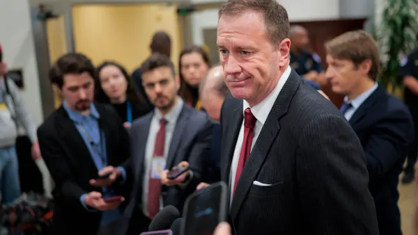 A person wearing a suit and tie talks into a microphone surrounded by other people.